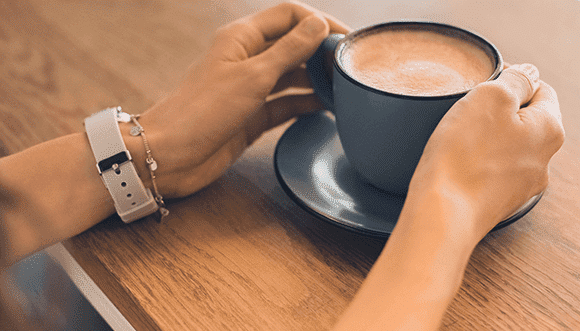 Hands holding a blue cup of coffee on a wooden table, showcasing a casual coffee moment. A light-colored bracelet and watch are visible on one wrist, adding a touch of style.