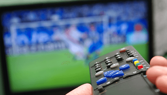 A close-up of a hand holding a TV remote control in front of a blurred television screen displaying a soccer match. The image highlights the engagement in sports viewing at home, showcasing the excitement of live sports events.