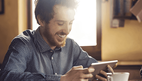 Smiling man using a tablet in a cozy café setting, enjoying his digital experience while seated at a table. Natural light illuminates the scene, creating a warm atmosphere.