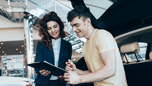 A smiling car salesperson reviews paperwork with a young customer inside a modern car dealership, showcasing a positive buying experience.
