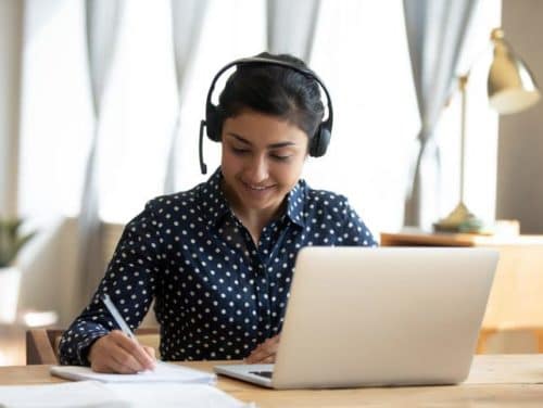 Woman wearing headphones, sitting at a desk with a laptop, taking notes on paper while engaged in online learning or virtual communication.
