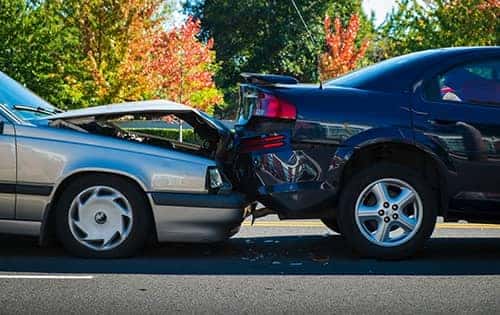 Image of a silver car with front-end damage colliding into the back of a black car, showcasing the aftermath of a rear-end traffic accident. Trees with autumn foliage in the background enhance the setting.