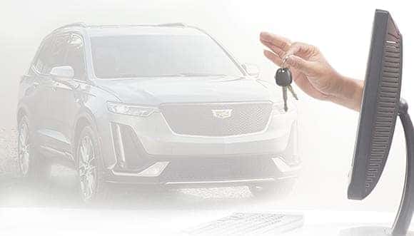 A hand holding car keys in front of a computer screen, with a luxury SUV in the background, illustrating online car buying and digital vehicle ownership.