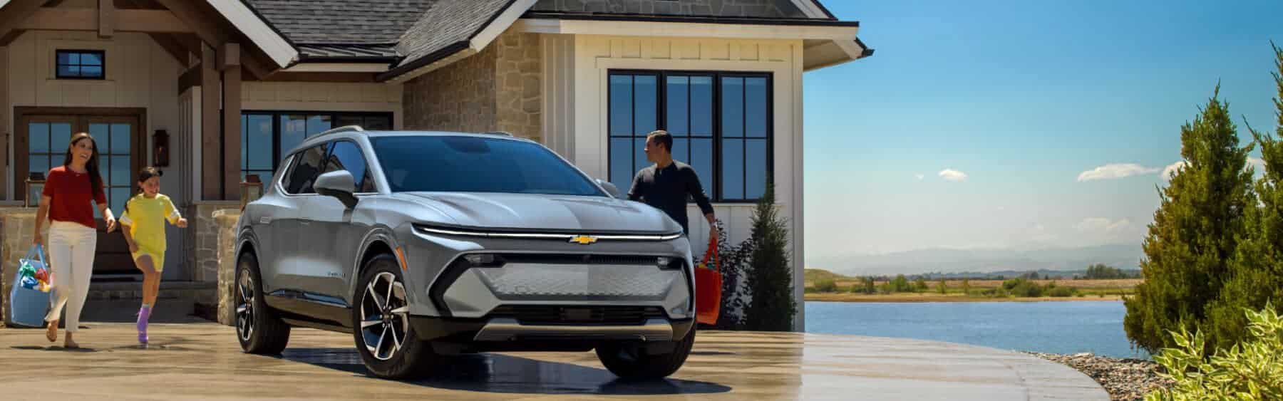 Family enjoying time together outside their home with a Chevrolet SUV parked in the driveway, showcasing modern design and a scenic backdrop of water and greenery.