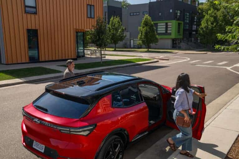 A vibrant red electric vehicle parked on a modern urban street, with two individuals approaching it near contemporary buildings surrounded by greenery. The scene highlights eco-friendly transportation and urban living.