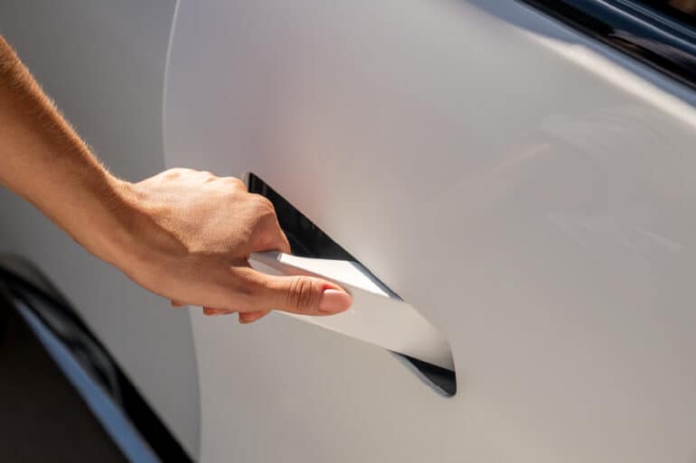 Close-up of a hand reaching for the handle of a white car door, illustrating the action of opening a vehicle. The focus is on the sleek design of the car's exterior and the hand's grip on the handle.