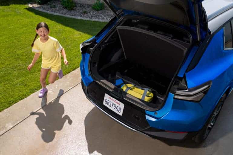 A young girl in a yellow outfit joyfully runs towards a blue SUV with its trunk open, revealing a yellow backpack and other items. The scene is set on a sunny day in a grassy yard, showcasing family-friendly vehicle features and outdoor fun.