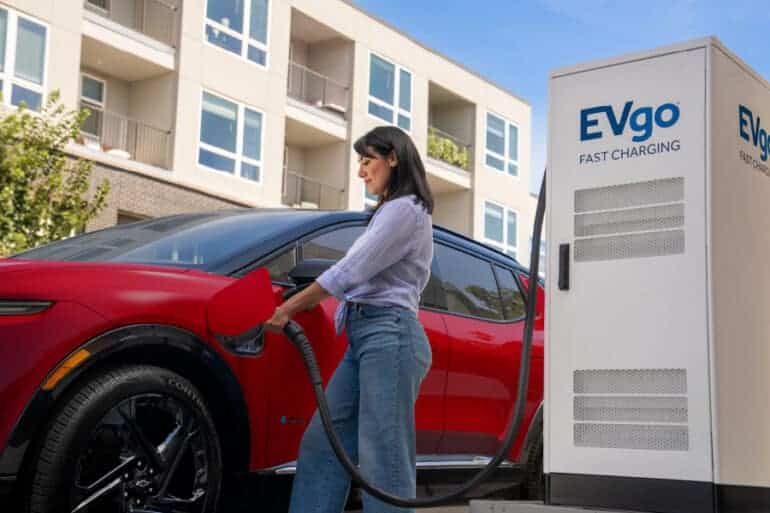 A woman charging a red electric vehicle at an EVgo fast charging station, with modern apartment buildings in the background. The image highlights the convenience of electric vehicle charging in urban settings.