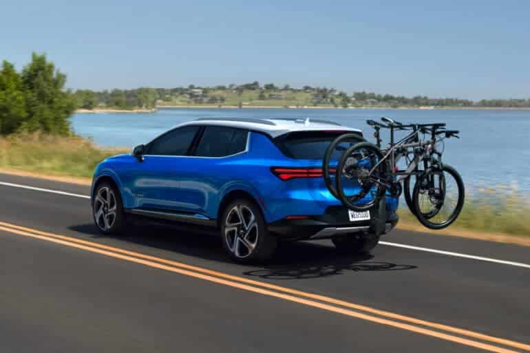 A blue SUV with bicycles mounted on a rear rack drives along a scenic lakeside road, showcasing an ideal setting for outdoor adventures and leisure activities.