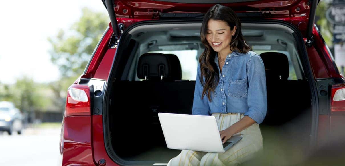 A young woman sitting in the back of a red SUV, smiling and working on a laptop, with trees and a sunny outdoor environment in the background.