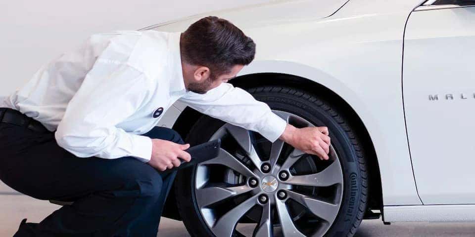 A mechanic inspects the tire of a white Chevrolet Malibu, using a tool to check for tread depth and wear, ensuring the vehicle's safety and performance.