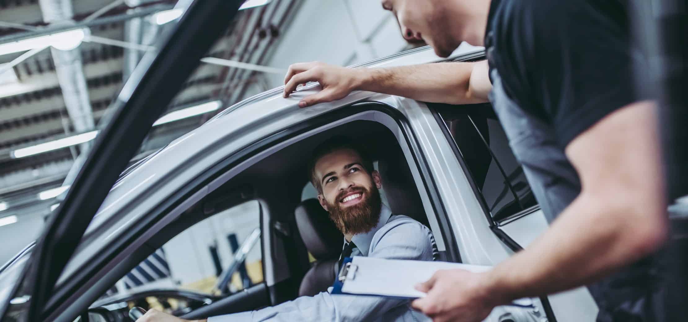A smiling customer and a mechanic interact in an auto repair shop as the mechanic holds a clipboard, discussing vehicle services.