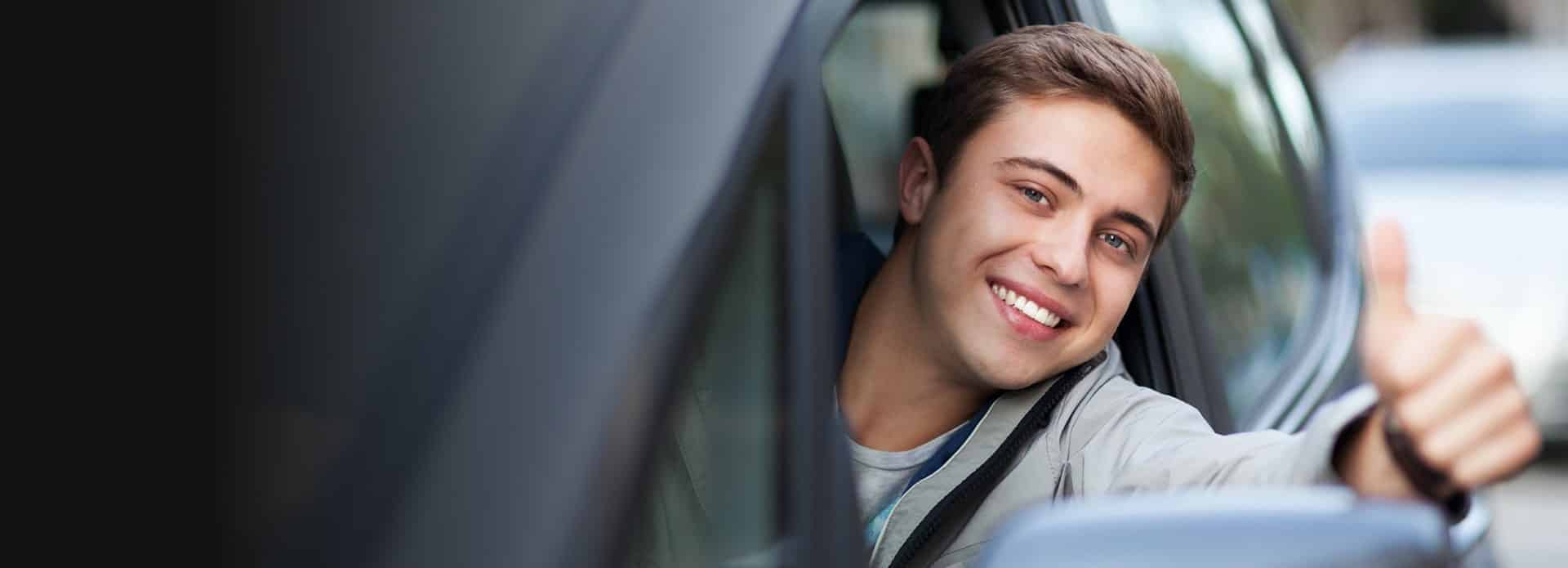 Smiling young man giving a thumbs up from the driver's seat of a car, representing positive driving experiences and customer satisfaction in car services.
