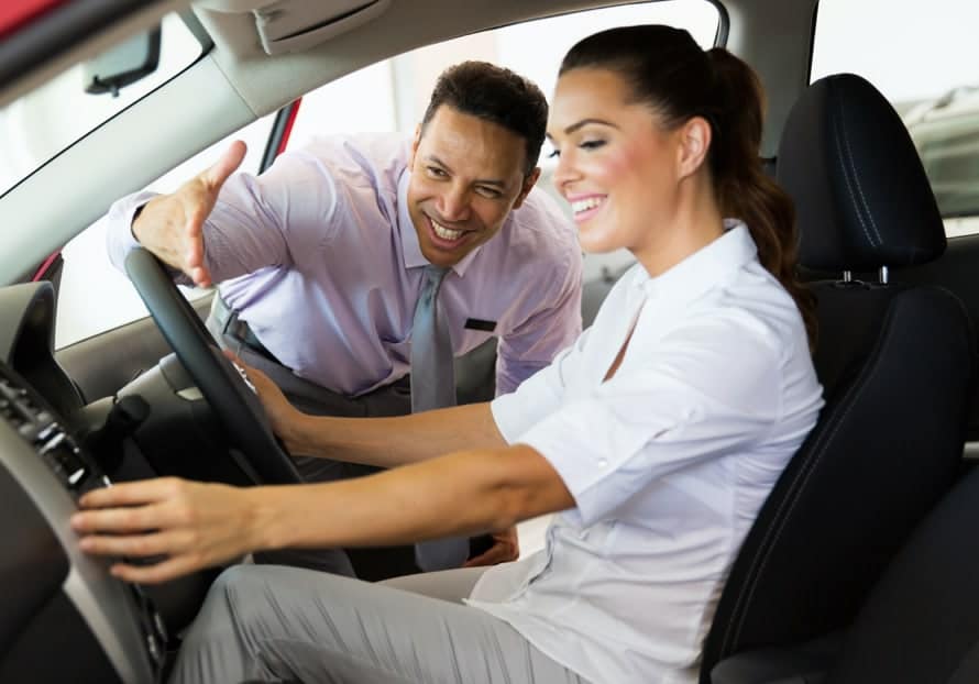 Car dealership scene with a smiling salesman gesturing towards a woman inside a new car, demonstrating features and providing guidance during a test drive.