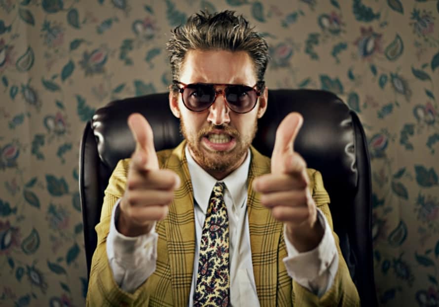 A man in a yellow plaid suit and sunglasses sits confidently in a leather chair, pointing his fingers like guns while giving an intense expression, set against a floral wallpaper background.