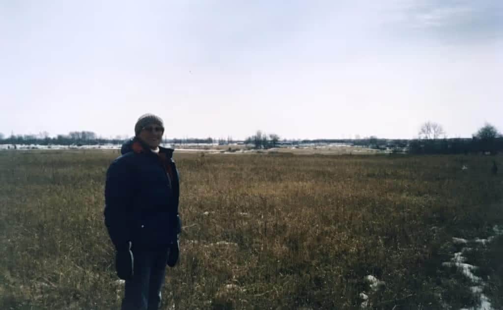 A person stands in a vast, winter landscape, surrounded by tall, dry grass and patches of snow under a clear, gray sky. The scene captures the serene beauty of nature during the colder months.