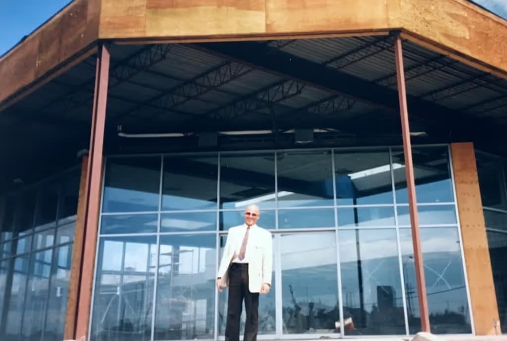 Architect standing in front of a modern, glass-fronted building with a rust-colored metal facade, showcasing contemporary architectural design against a clear blue sky.