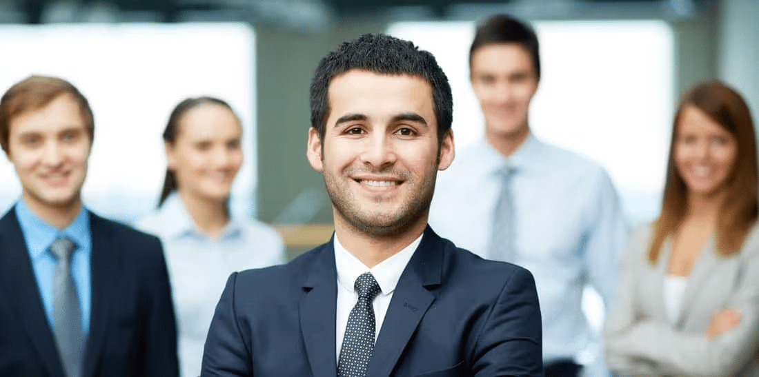 Group of diverse professionals smiling in a modern office setting, with a confident man in a suit in the foreground, showcasing teamwork and collaboration in a business environment.