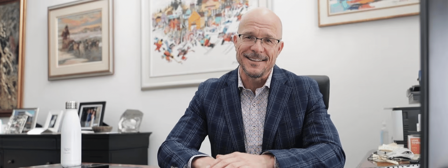 Professional man in a checkered blazer seated at his desk, smiling with a white water bottle and framed photos in the office background. Artworks adorn the walls, showcasing a vibrant and inviting workspace.